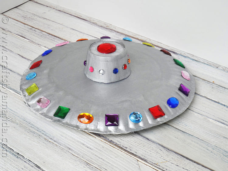 paper plate craft flying saucer with gems of different colors on wooden background