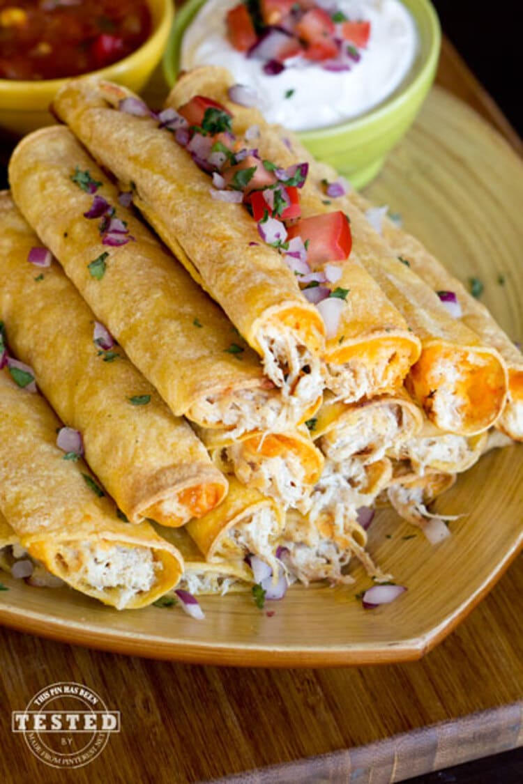 Crockpot Cream Cheese Taquitos - pile of tightly rolled up cheese and chicken taquitos with sour cream and salsa dip on the side