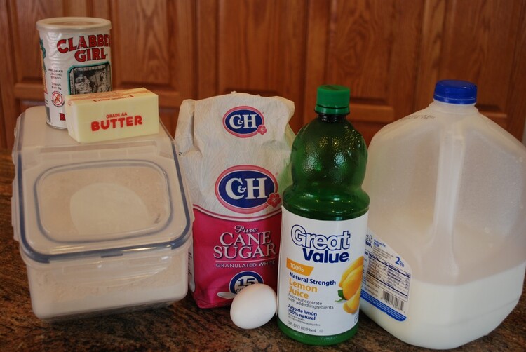 ingredient list on a worktop: Flour tub, with butter stick and baking powder on top, bag of cane sure, 1 egg and lemon juice bottle and a large bottle of milk.