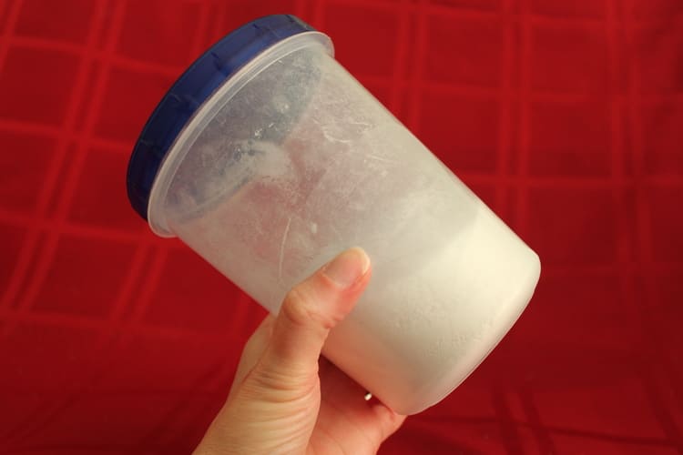 DIY make your own dishwasher detergent - homemade dishwasher detergent in a plastic container with a blue cup and a hand holding it on a red background