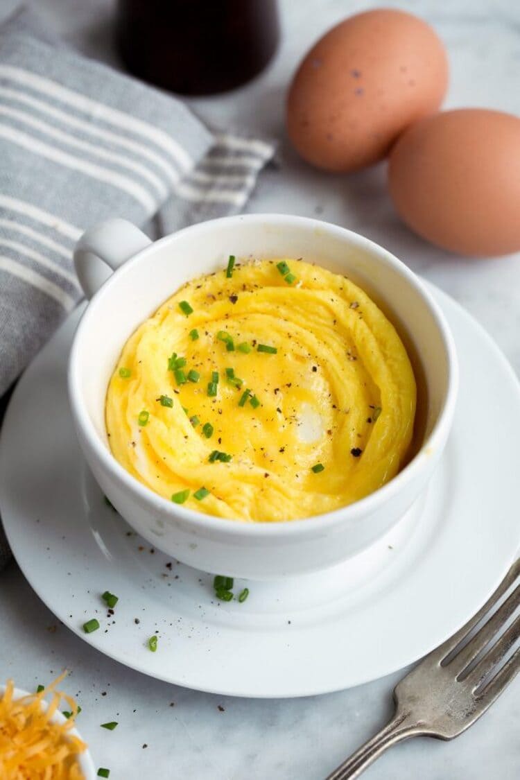 microwave mug snacks scrambled eggs in a white cup with a fork next to it, eggs behind it and a grey kitchen towel