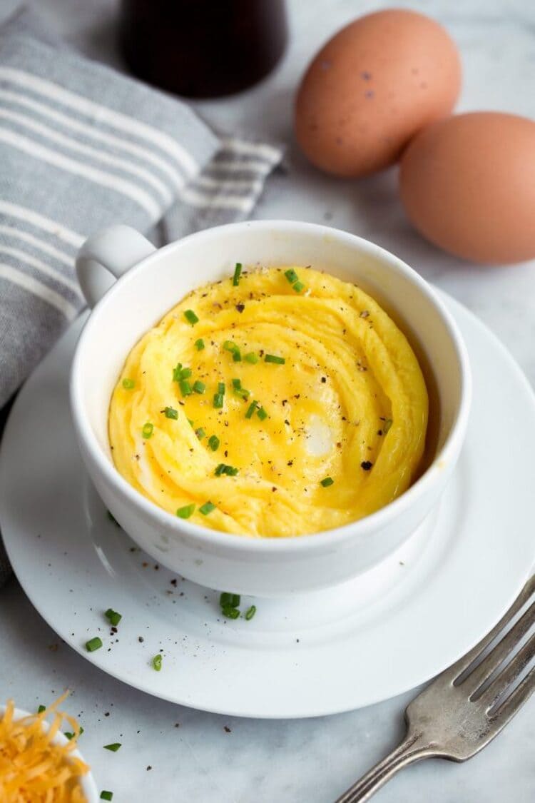 microwave mug snacks scrambled eggs in a white cup with a fork next to it, eggs behind it and a grey kitchen towel